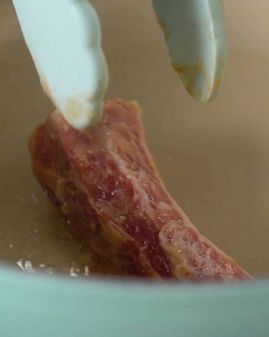 Une pince déposant un morceau de travers de porc mariné dans une cocotte bleu clair chauffée.
