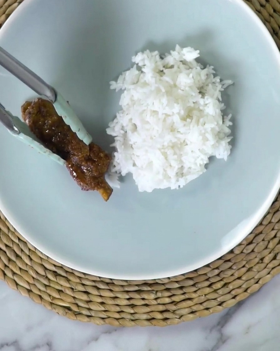 Une pince à embout silicone déposant un travers de porc glacé et collant sur une assiette bleu clair à côté de riz blanc moelleux.
