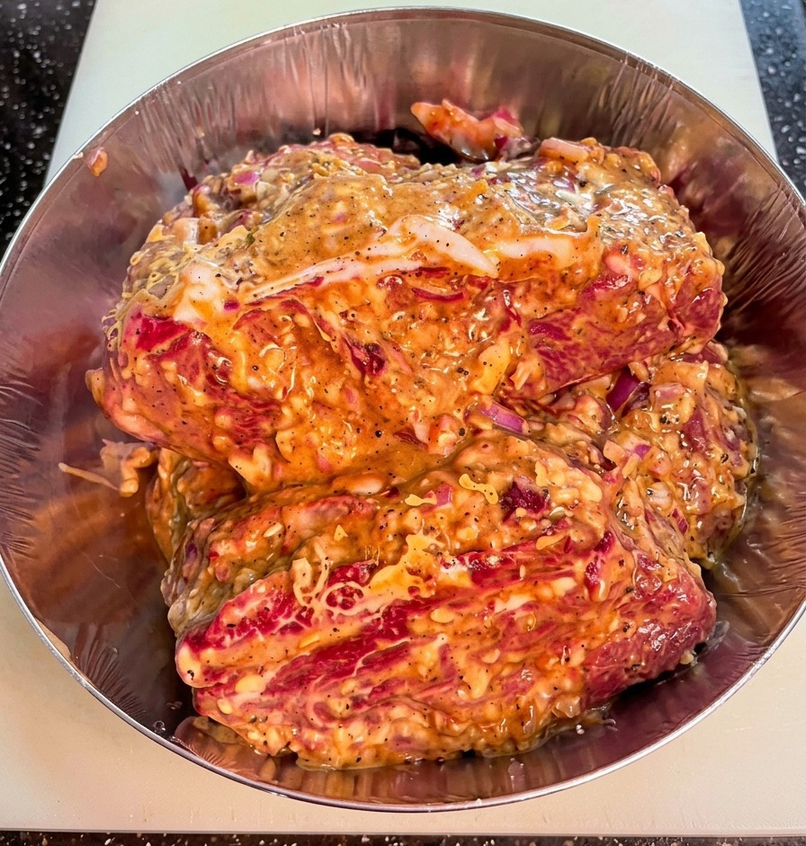Gros plan de morceaux de bœuf dans un bol en métal, enrobés dune marinade épaisse avec des éclats doignon et de poivre noir.