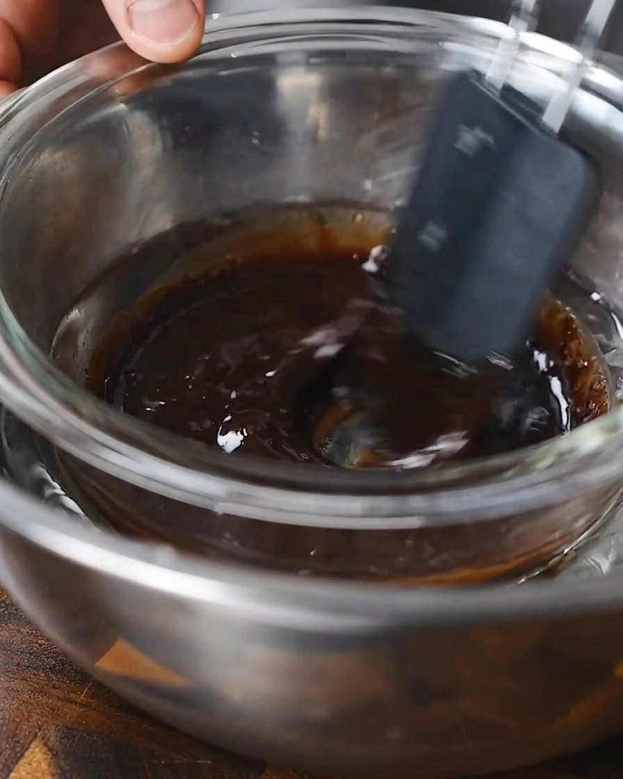 Une spatule remuant une marinade liquide sombre dans un bol en verre placé dans un bain-marie.