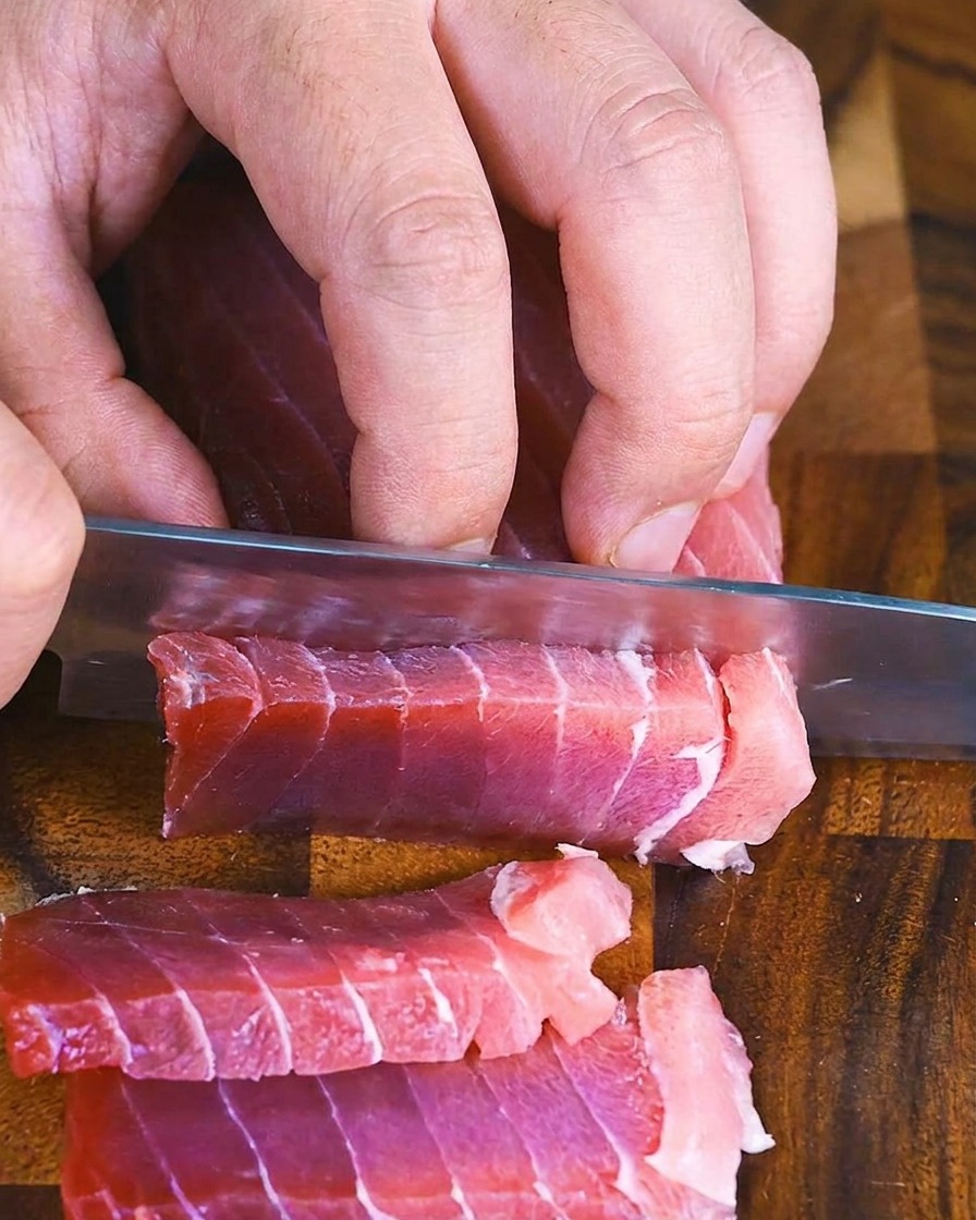 Mains utilisant un couteau de chef pour trancher un bloc de thon rouge cru en rectangles épais et réguliers.