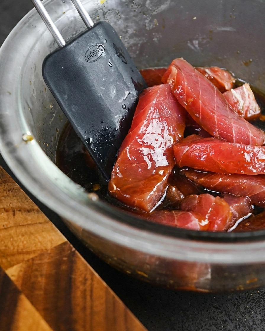 Tranches de thon rouge cru mélangées dans un bol de marinade liquide sombre avec une spatule.