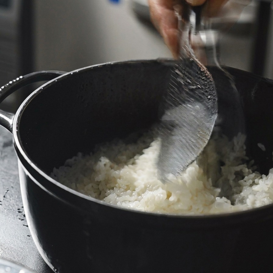 Une main utilisant une spatule à riz transparente pour remuer et servir du riz blanc fumant dans une casserole noire.