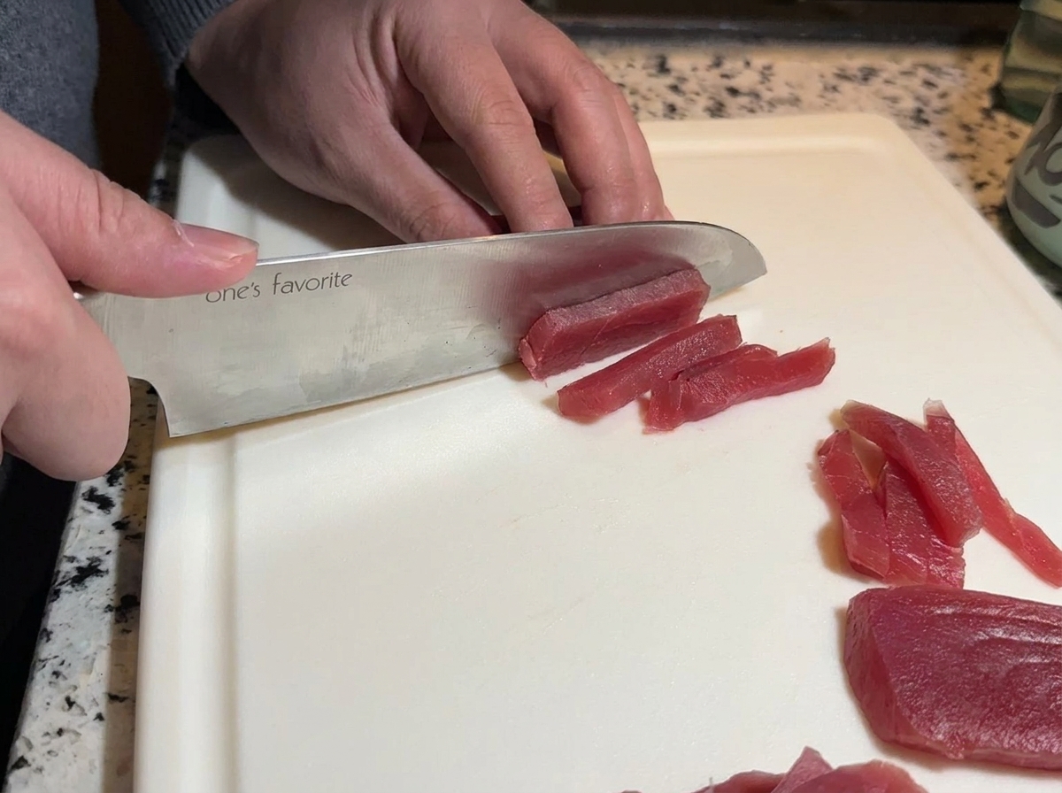 Un couteau de chef tranchant découpe un pavé de thon rouge cru en longues lanières.