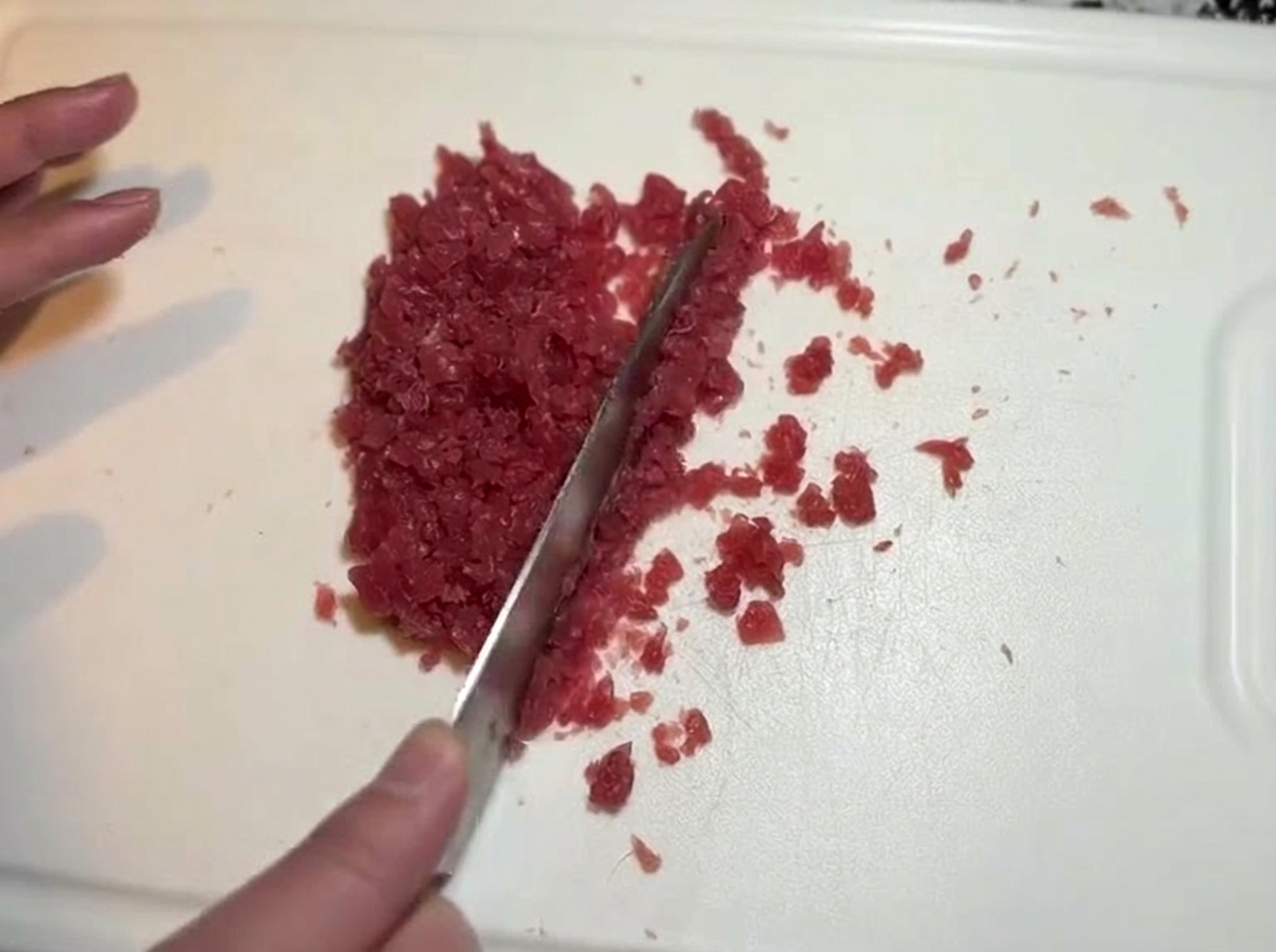 Un couteau hache finement des morceaux de thon rouge cru en une texture de pâte sur une planche.
