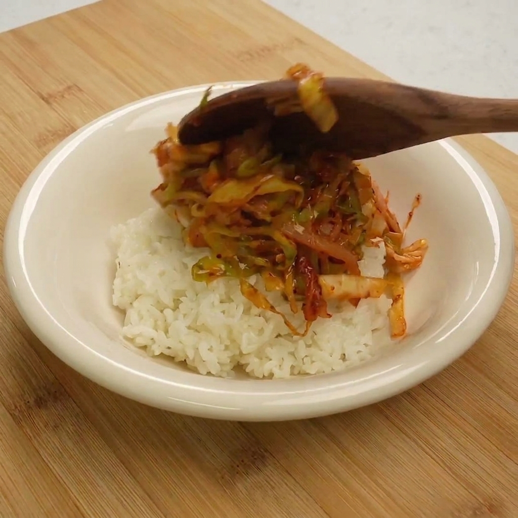 Une cuillère en bois servant le sauté de thon et chou sur un monticule de riz blanc dans un bol.