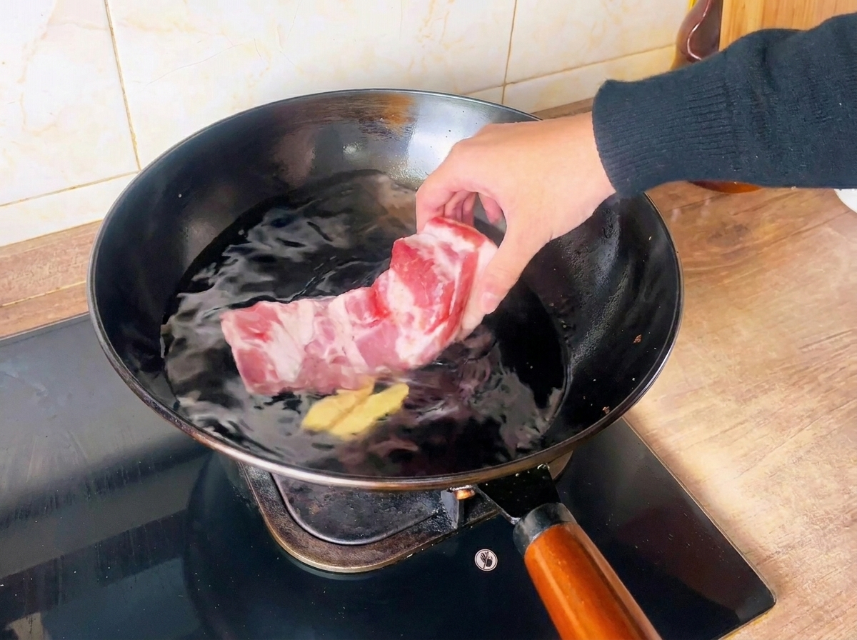 Une main place un bloc de poitrine de porc crue dans un wok rempli deau froide et de tranches de gingembre.