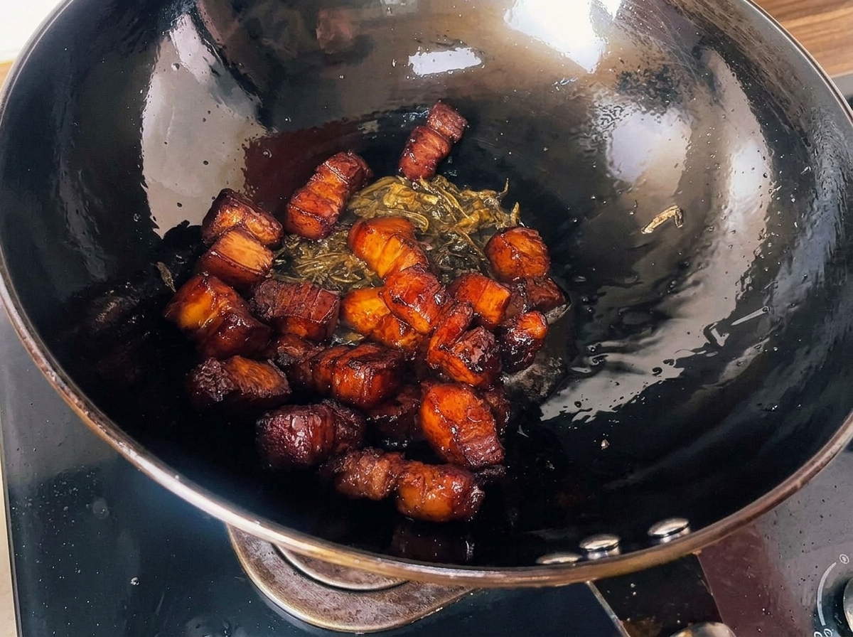 Cubes de porc rôtis au glaçage sombre mélangés avec des feuilles de thé frites croustillantes à lintérieur dun grand wok noir.