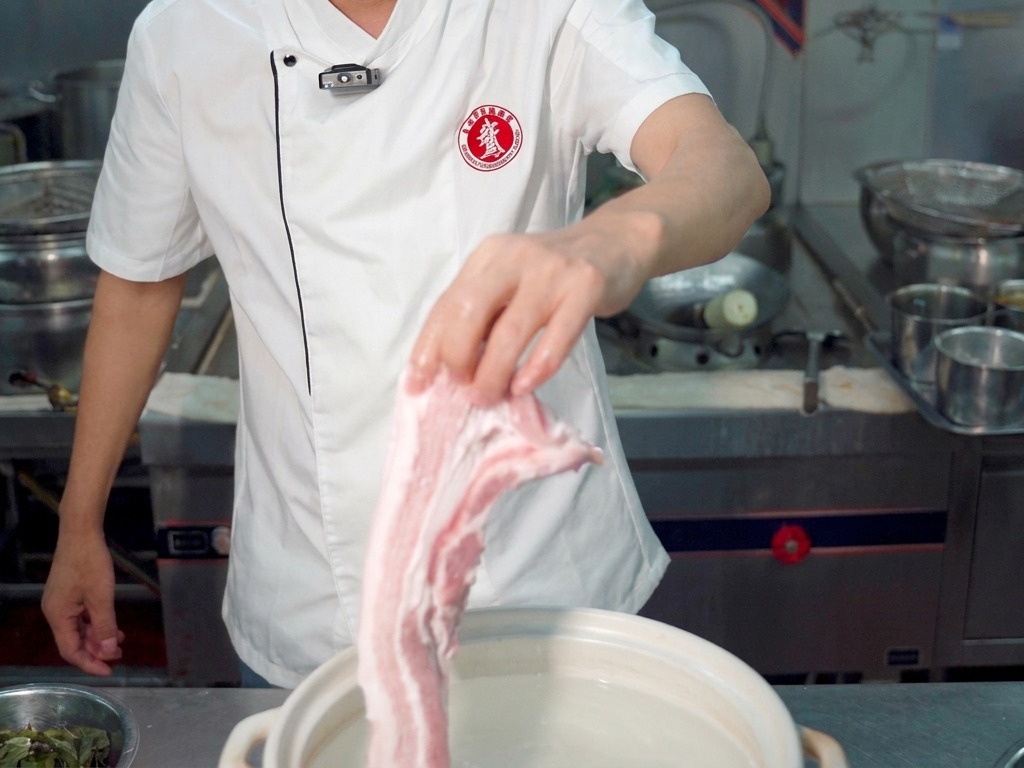 Un chef tenant un gros morceau de poitrine de porc crue au-dessus dune marmite blanche remplie deau.