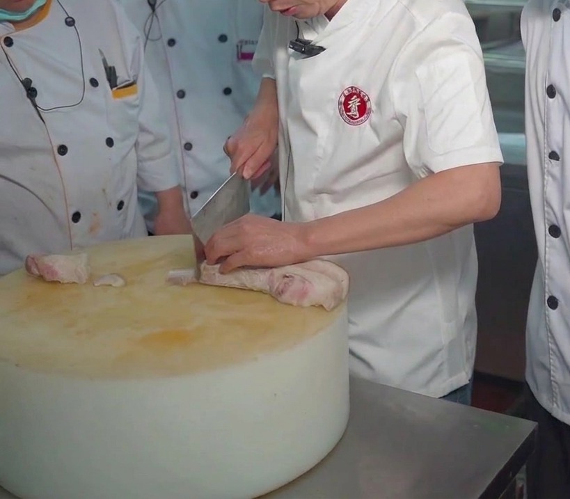 Un chef utilisant un couperet pour trancher un bloc de poitrine de porc bouillie sur une grande planche à découper ronde et épaisse.