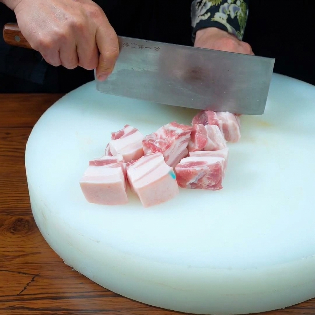 Mains utilisant un couperet pour trancher une pièce de porc à trois couches en cubes uniformes sur une planche à découper blanche.