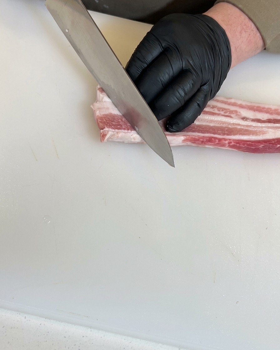 Mains gantées utilisant un couteau de chef pour effectuer des incisions diagonales profondes dans un morceau de poitrine de porc crue sur une planche à découper blanche.