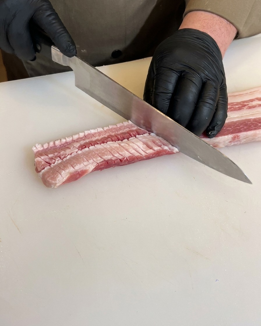 Mains gantées incisant lenvers dune poitrine de porc crue avec un couteau, montrant des coupes en damier profondes sur la viande.
