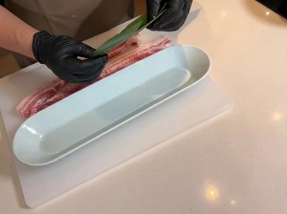 Mains tenant une feuille de bambou verte au-dessus dun long plateau de service blanc, avec la poitrine de porc incisée reposant à proximité sur la planche.