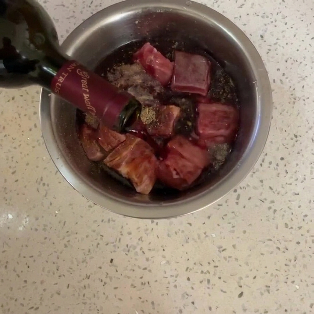 Vin rouge versé depuis une bouteille en verre foncé sur le porc dans un bol en inox.