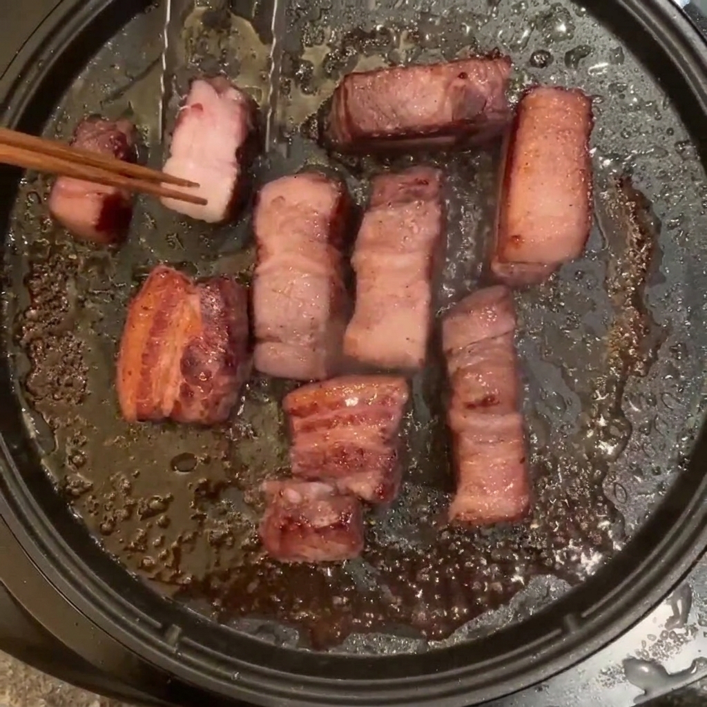 Morceaux de porc frits dans une poêle électrique, retournés avec des baguettes en bois.