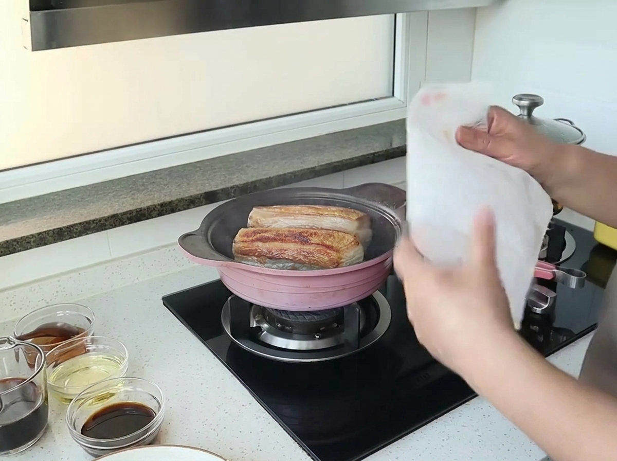 Mains tenant un essuie-tout blanc au-dessus dune poêle avec deux morceaux de porc en train de saisir.