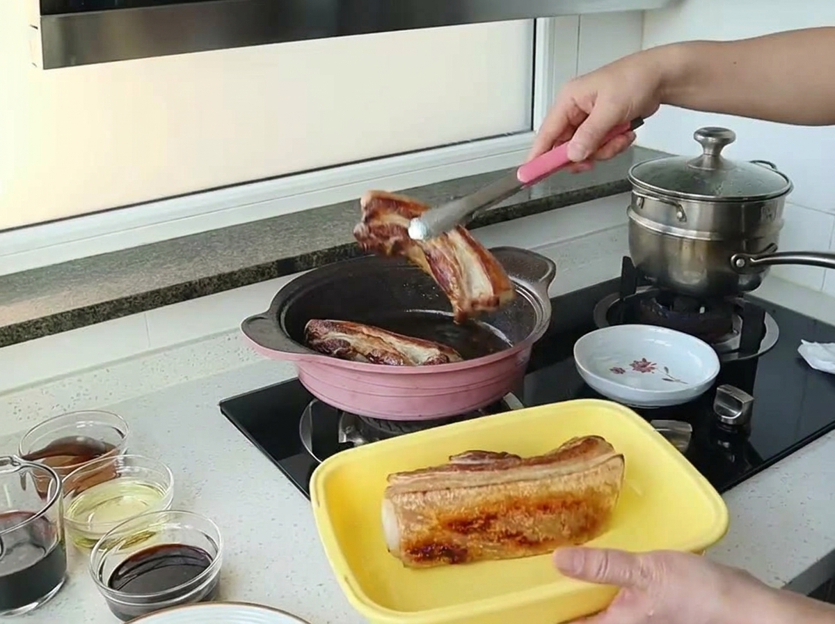 Pinces soulevant un morceau de poitrine de porc magnifiquement doré hors dune poêle rose.
