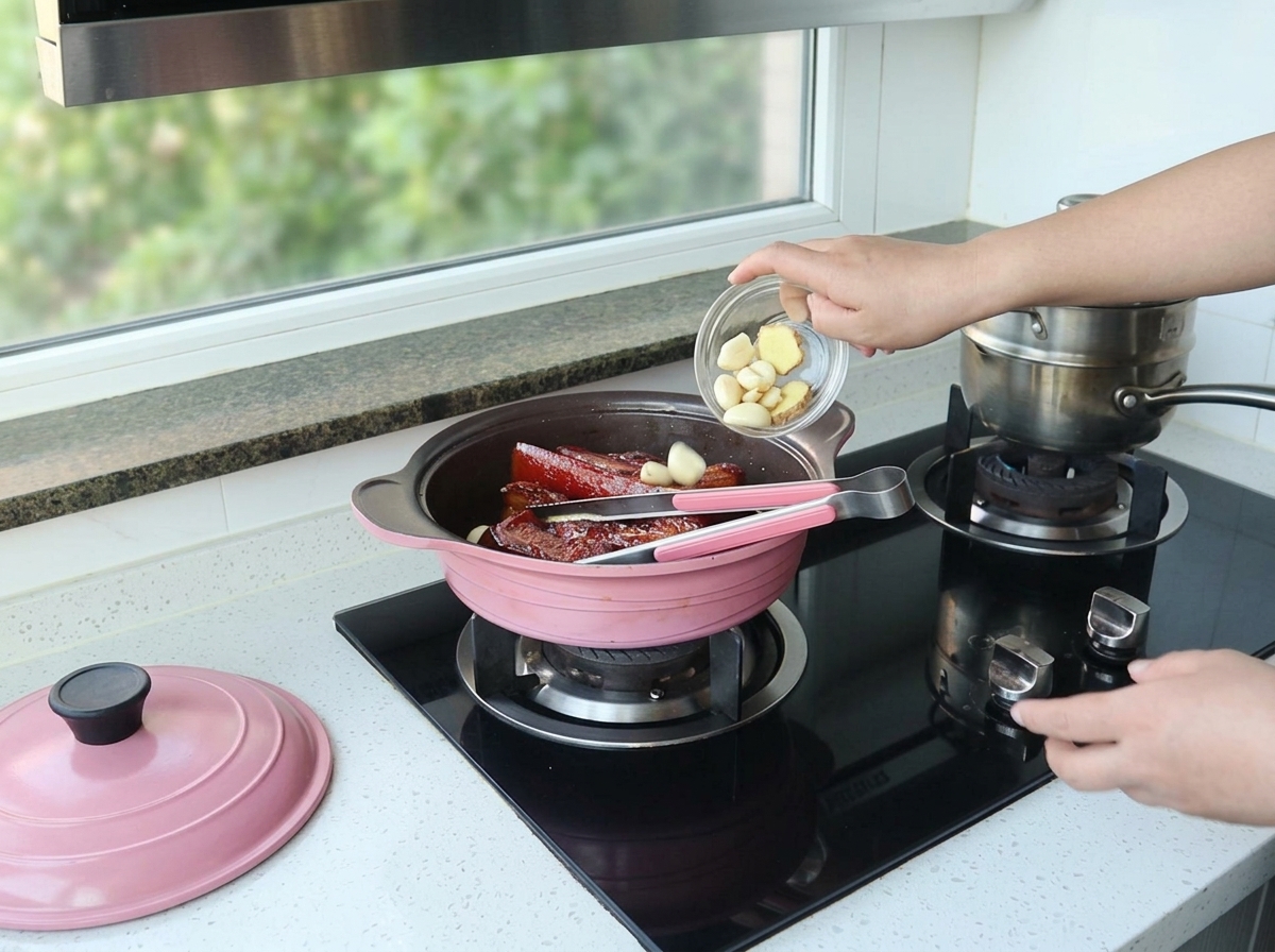 Main versant des gousses dail pelées dans une cocotte rose contenant le porc braisé.