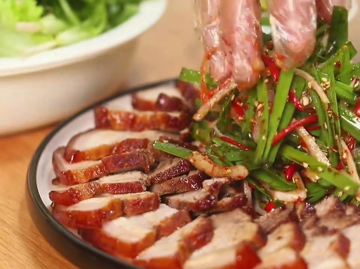 Mains gantées mélangeant une salade doignons nouveaux et piments à côté de tranches de porc laqué.