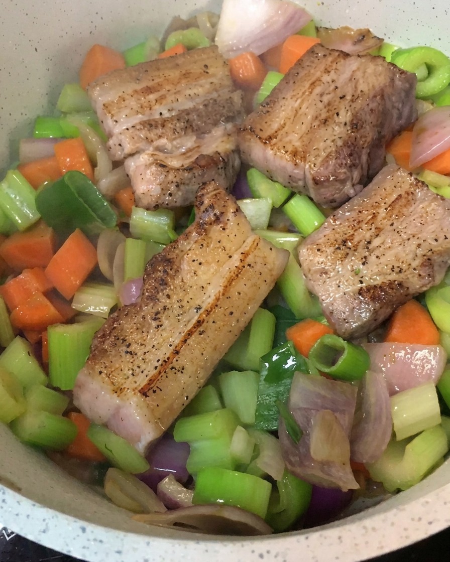 Morceaux de poitrine de porc saisis placés sur un lit de légumes sautés dans une marmite.