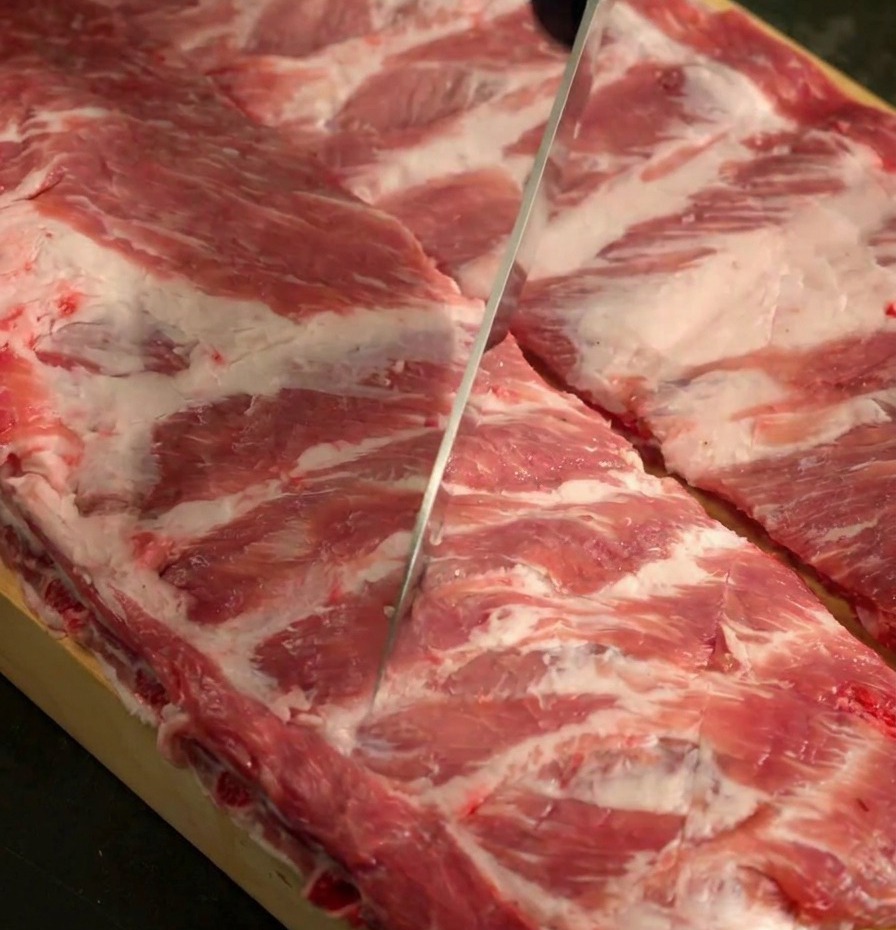 Un couteau aiguisé quadrillant la surface de deux racks de travers de porc crus sur une planche à découper.