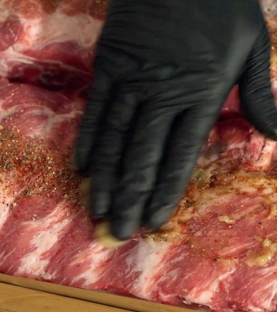 Une main gantée frottant les épices et lail sur des travers de porc crus.