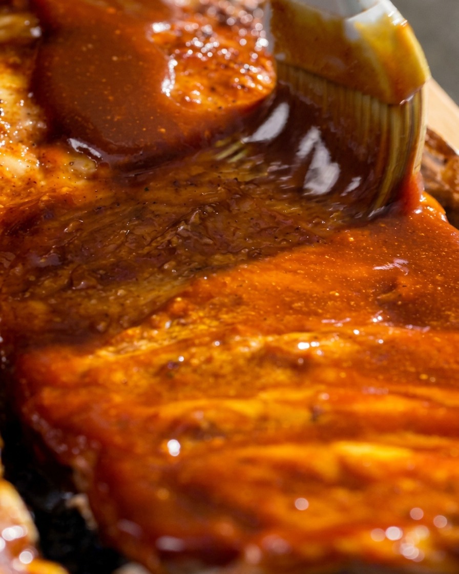 Gros plan dun pinceau étalant un glaçage barbecue épais et brillant sur les travers de porc cuits.