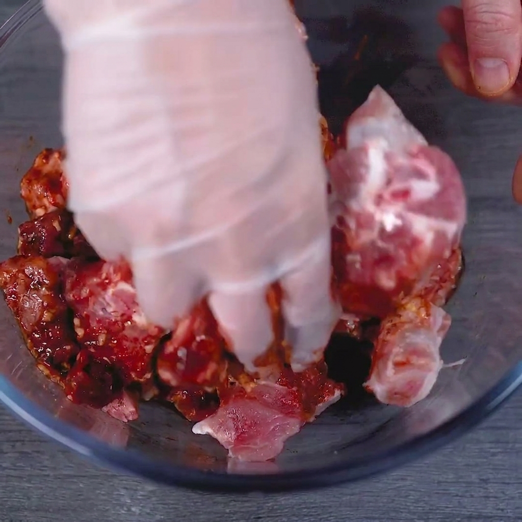 Une main gantée massant vigoureusement la marinade rouge sur les travers de porc dans un bol en verre.