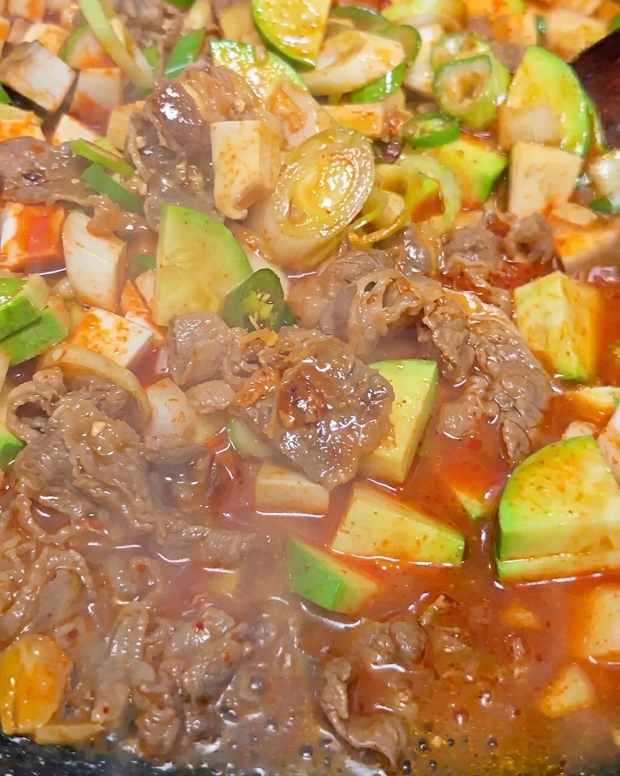 Gros plan sur le ragoût coréen à la pâte de soja bouillonnant intensément, montrant des morceaux de tofu, de bœuf et de courgette verte dans un bouillon brun rougeâtre.