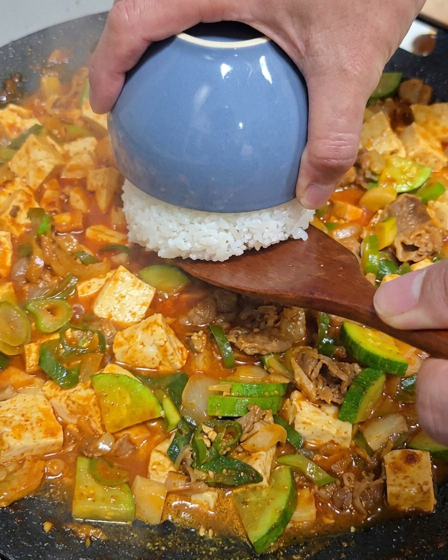Une main utilisant une spatule en bois pour placer un petit bol bleu de riz blanc à lenvers au centre de la poêle remplie de ragoût de bœuf et de légumes.
