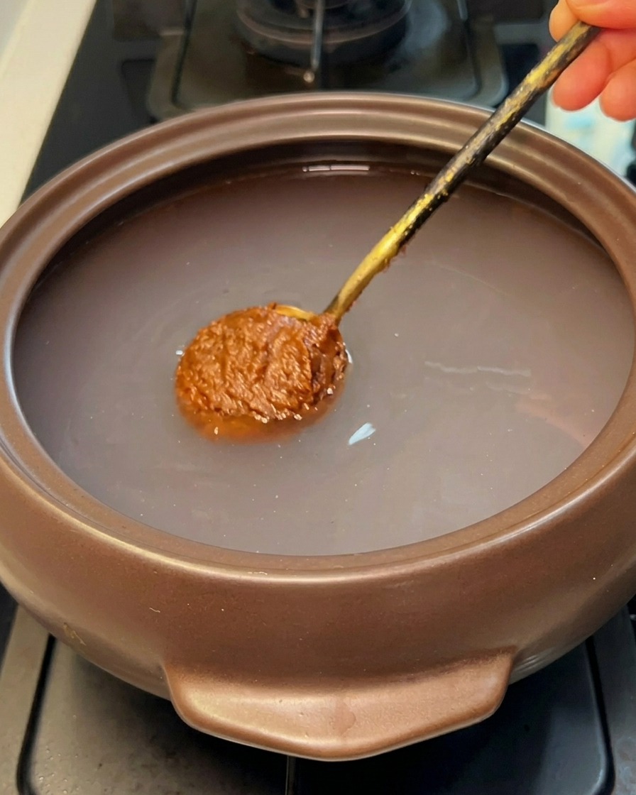 Une cuillerée de pâte de soja coréenne brune plongée dans leau claire dun pot en pierre.