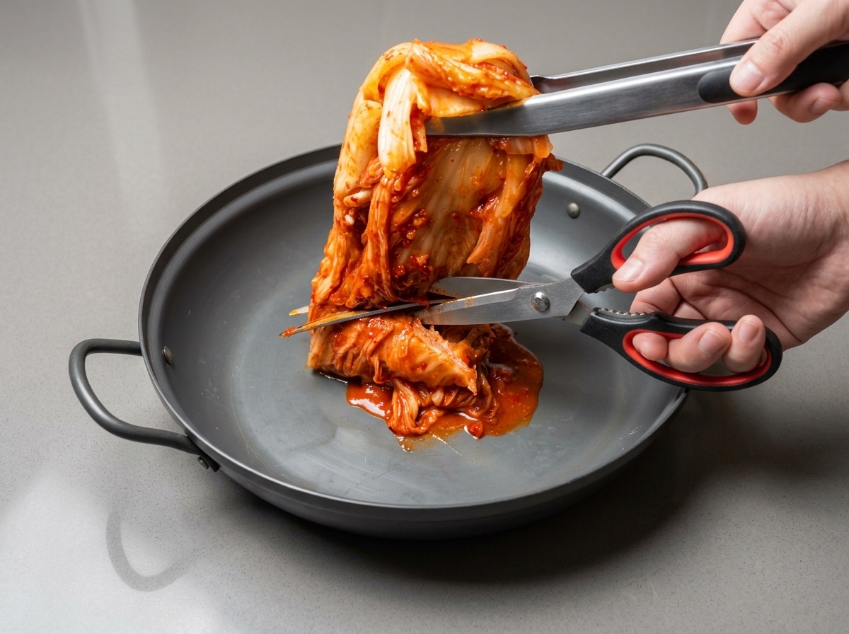 Des ciseaux de cuisine coupent un morceau de kimchi entier dans une grande sauteuse.