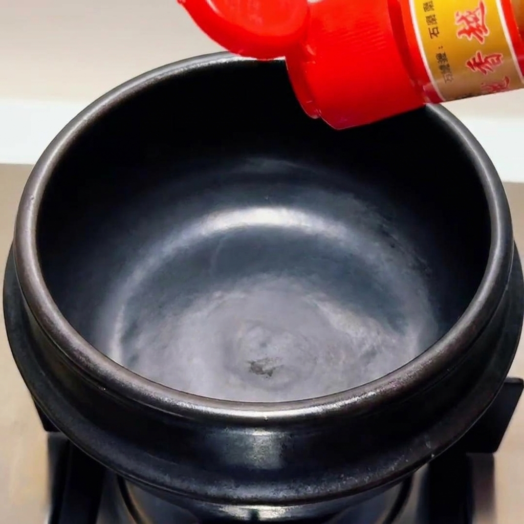 Huile de sésame versée dune bouteille à bouchon rouge dans un pot en terre cuite noir vide.