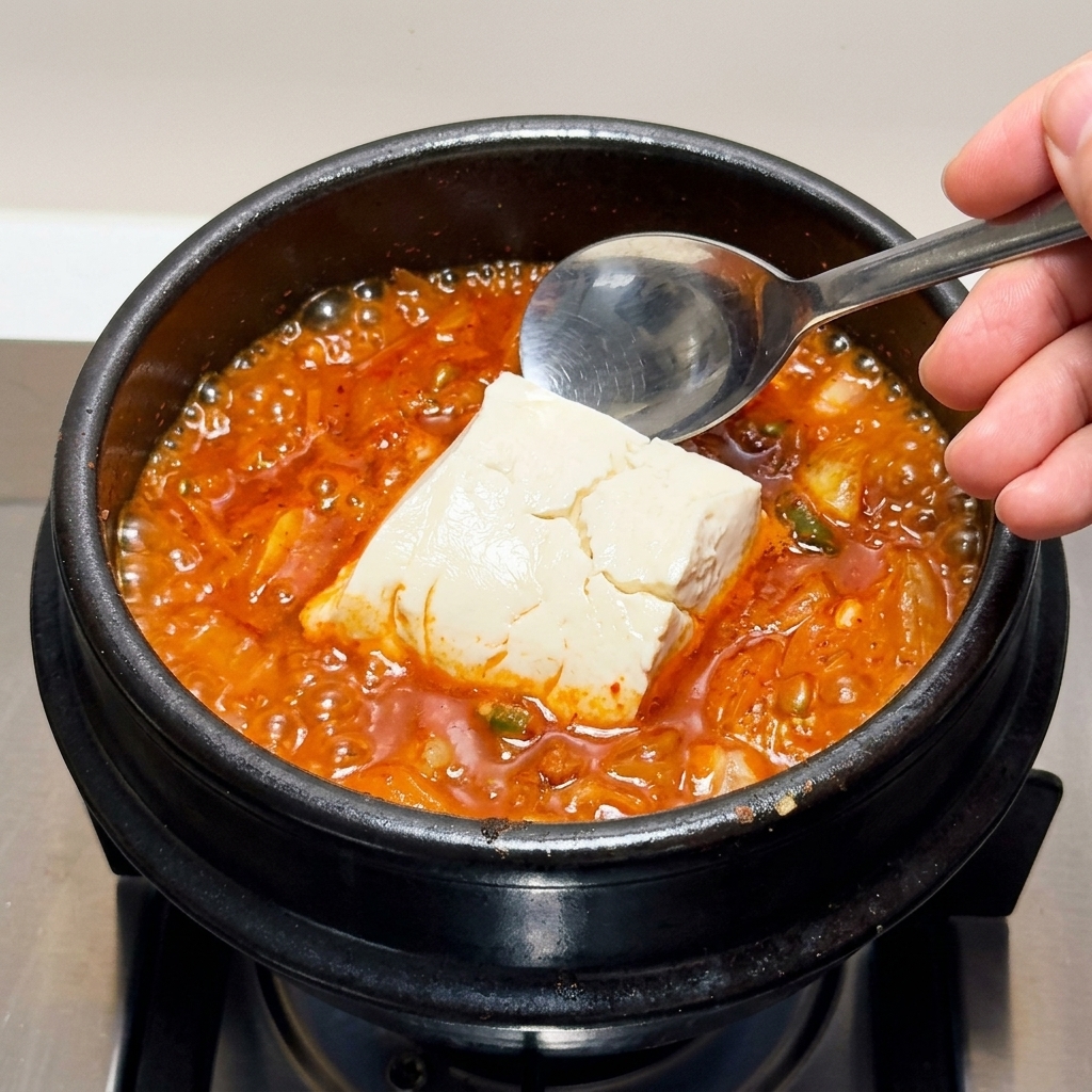 Gros morceaux de tofu soyeux soigneusement ajoutés à la louche dans le ragoût de kimchi bouillonnant.