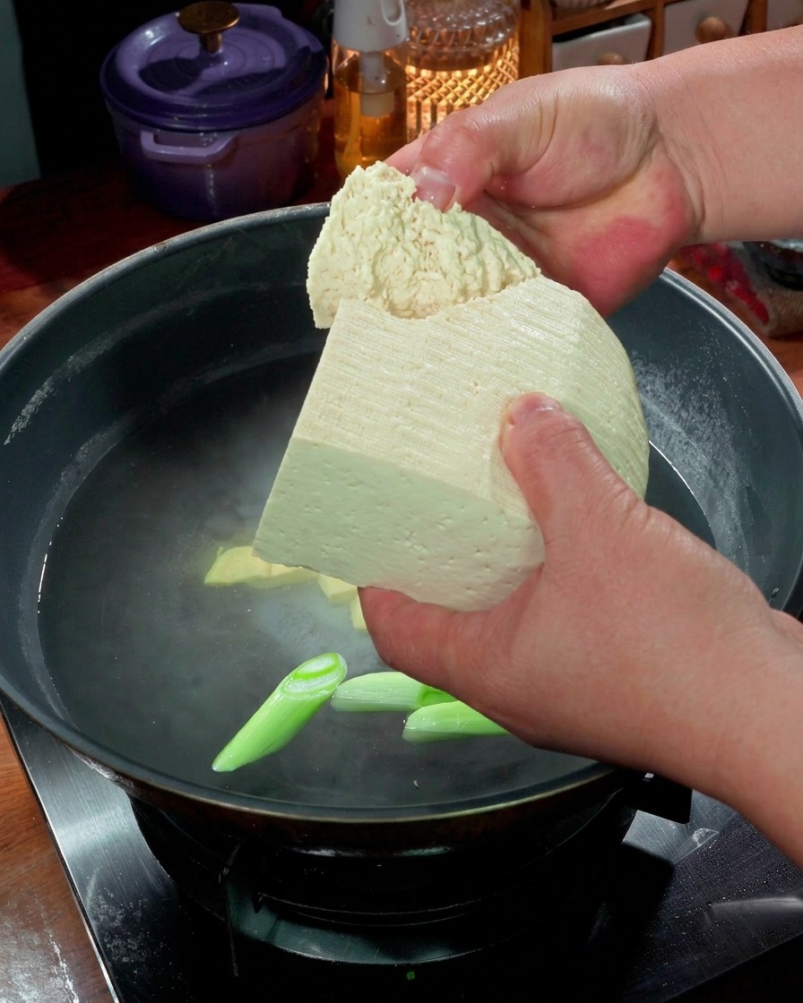 Des mains cassant de gros morceaux de tofu ferme dans une casserole deau frémissante avec des oignons verts.