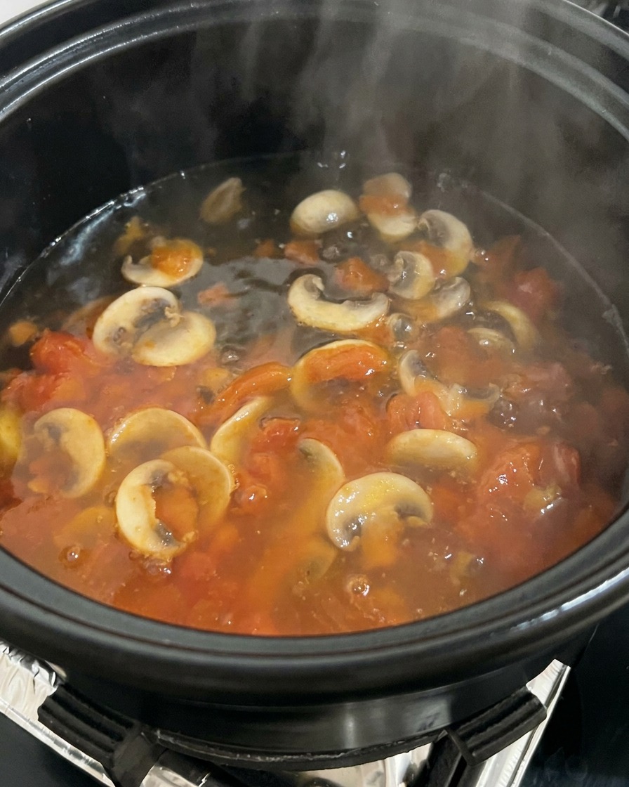 Eau bouillante versée dans une marmite remplie de tomates et de champignons, créant de la vapeur.