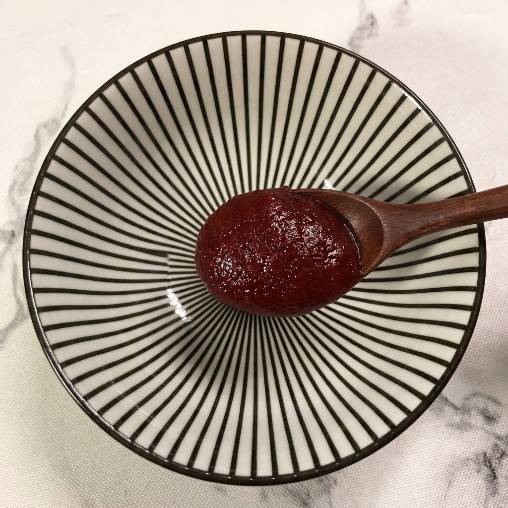 Une cuillère en bois ajoutant une généreuse cuillerée de pâte de piment coréenne dans un bol à motifs rayés.