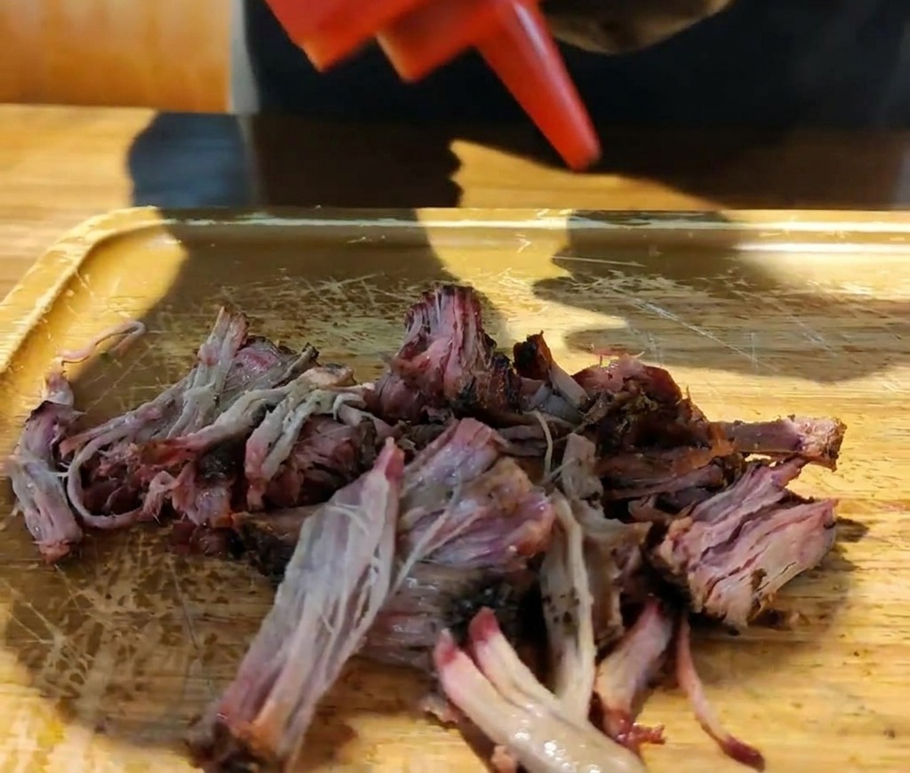 Une bouteille verse une sauce barbecue épaisse sur un tas de porc fumé effiloché sur une planche en bois.