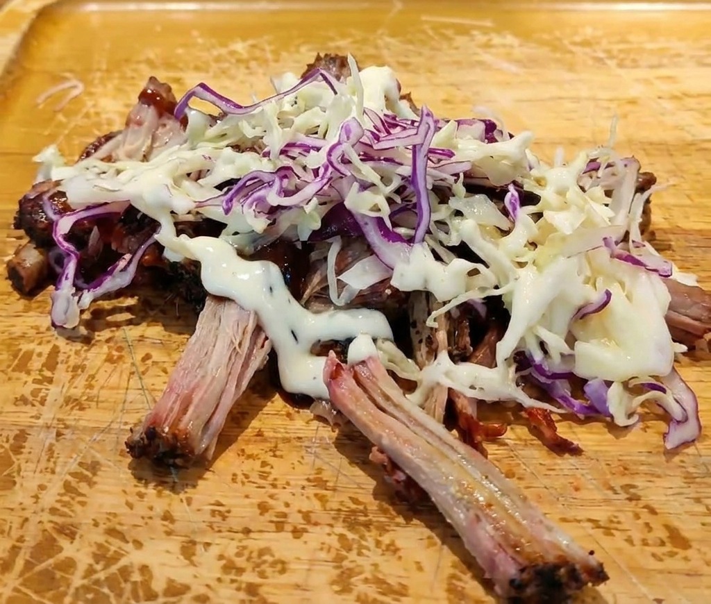 Porc effiloché à la sauce BBQ surmonté de coleslaw frais avec un filet de sauce crémeuse blanche sur une planche à découper.