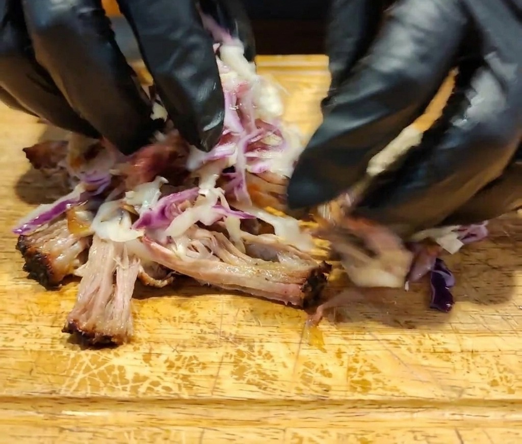 Des mains gantées mélangent la viande effilochée, la sauce foncée et le coleslaw coloré sur une surface en bois.