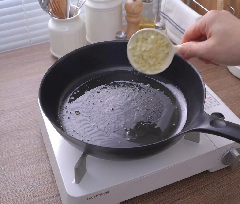 Une petite tasse blanche versant de lail haché dans une poêle contenant du beurre fondu.