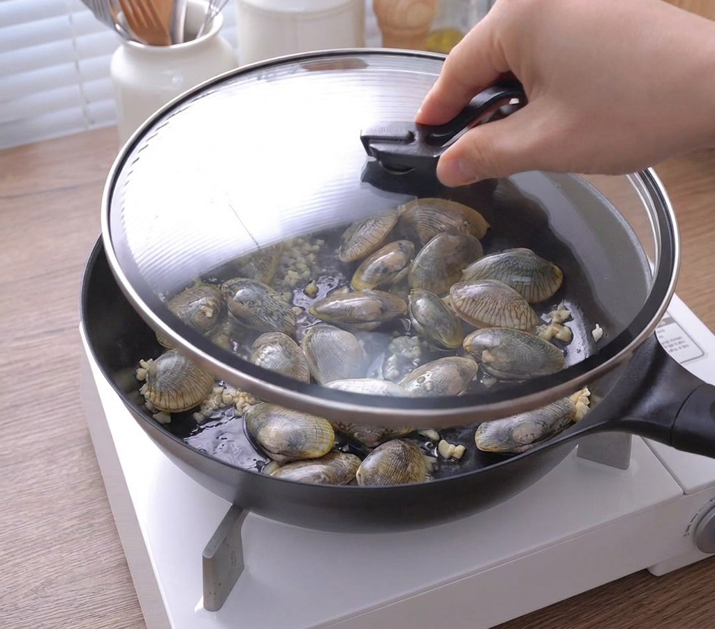Une main posant un couvercle en verre sur une poêle remplie de palourdes et dail au beurre.