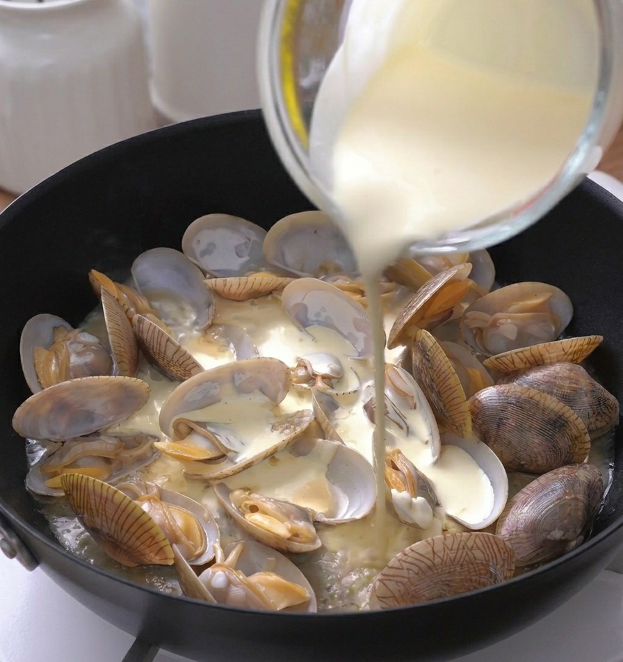 Crème liquide épaisse versée dun doseur en verre dans une poêle remplie de palourdes ouvertes.