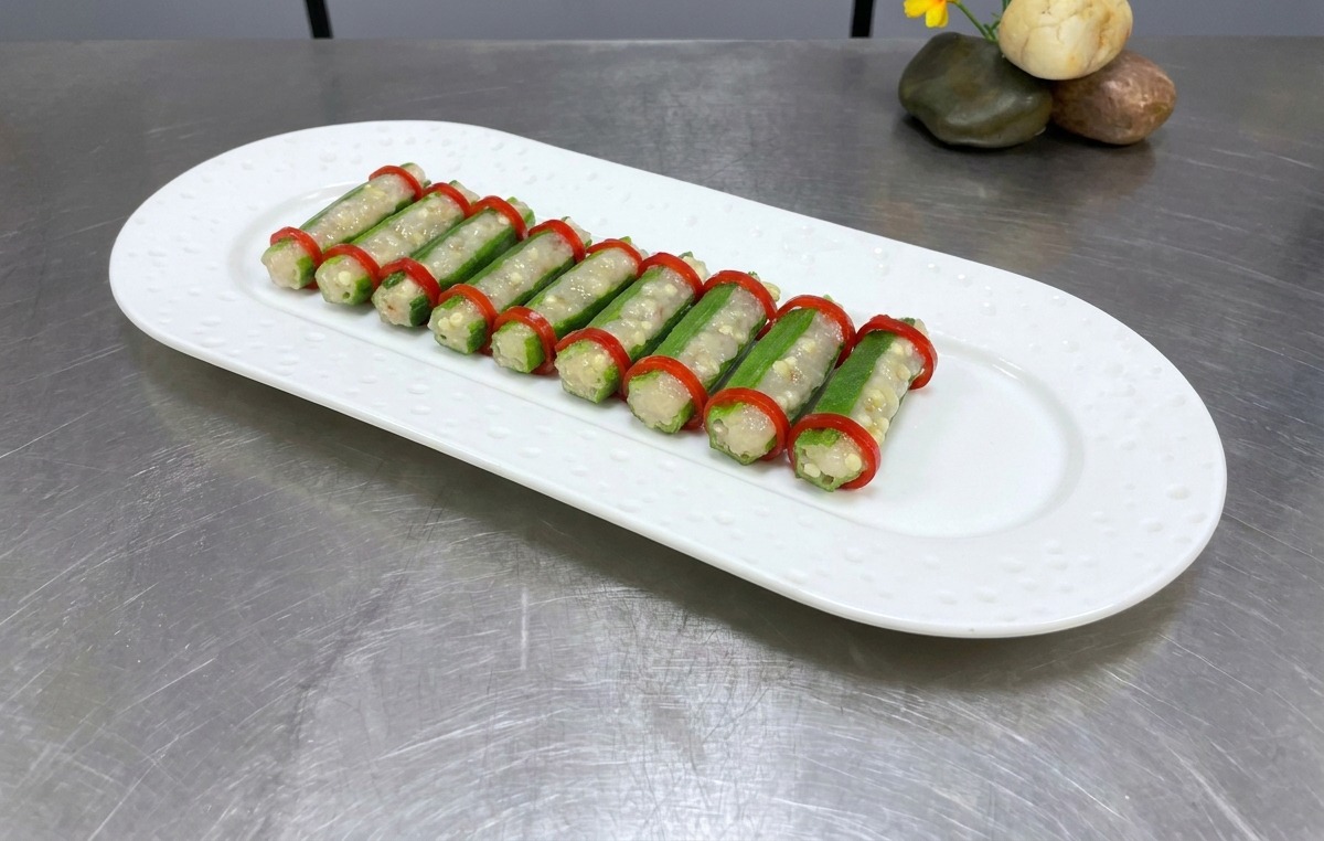 Deux rangées de gombos farcis avec des rondelles de piment sur une assiette ovale blanche, prêts pour la vapeur.
