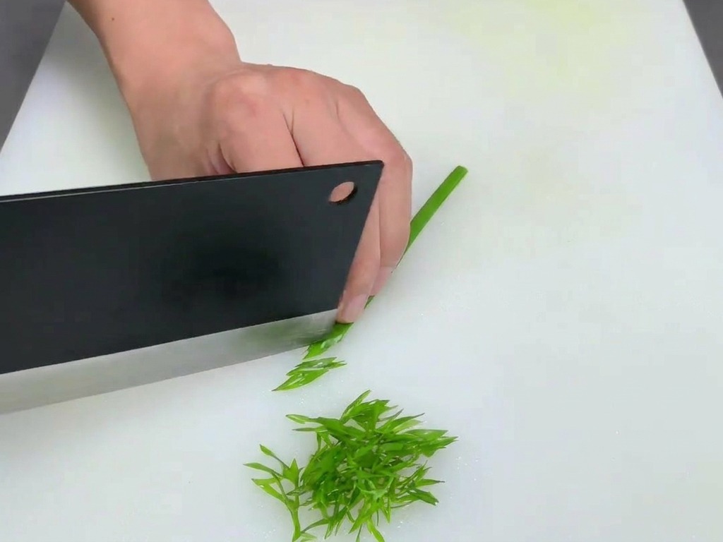 Une personne utilisant un grand couperet noir pour émincer finement des tiges doignons verts sur une planche blanche.