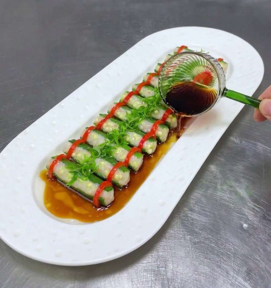 Une louche en verre versant de la sauce soja sur les gombos farcis et les oignons verts émincés sur une assiette blanche.