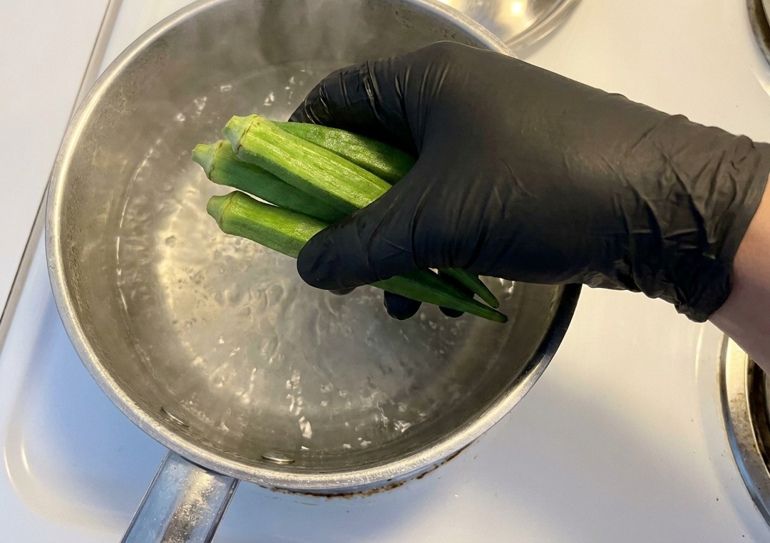 Une main gantée plongeant des gousses de gombo vert entières dans une casserole deau bouillante sur une cuisinière blanche.