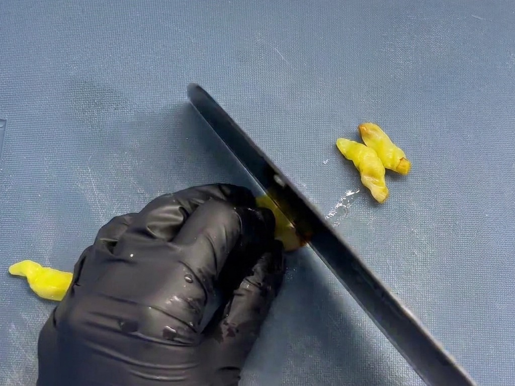 Un chef utilisant un couteau noir pour émincer finement des petits piments jaunes au vinaigre sur une planche à découper bleue.