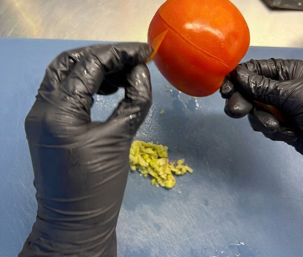 Mains en gants noirs retirant la peau fine dune grosse tomate rouge mûre au-dessus dun plan de travail.